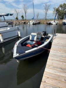 Small blue and white motorboat tied to a wooden dock in a calm marina, with other boats and sailboats in the background.